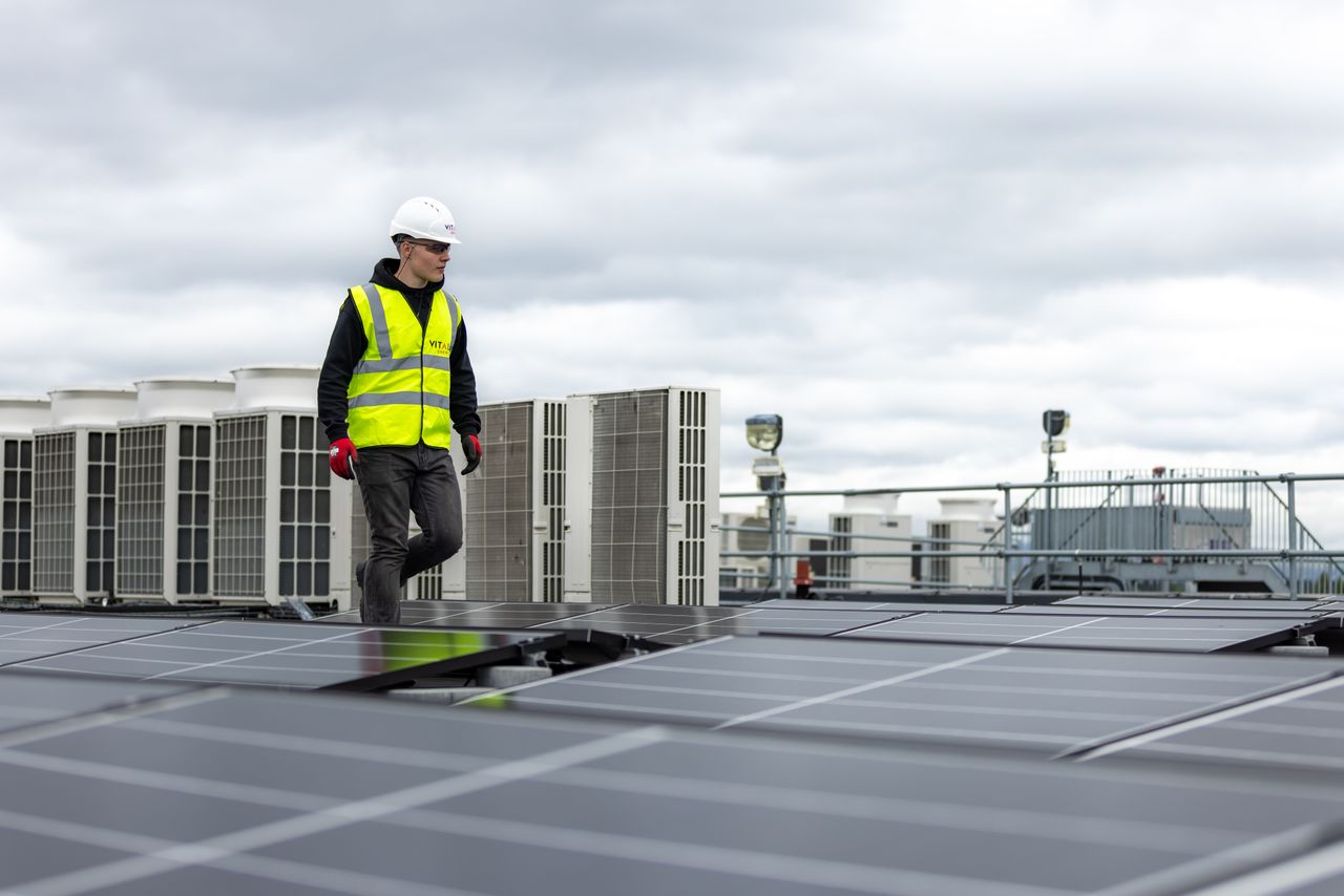 Engineer in safety gear inspecting solar panels on a rooftop under a cloudy sky.